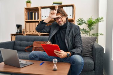 Handsome middle age man working at consultation office making fun of people with fingers on forehead doing loser gesture mocking and insulting. 