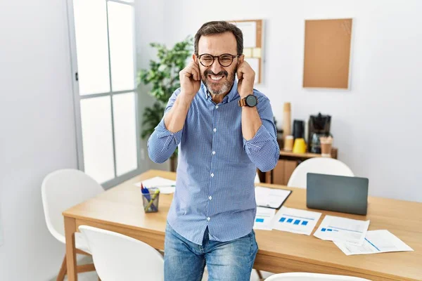 Middle age hispanic man with beard wearing business clothes at the office covering ears with fingers with annoyed expression for the noise of loud music. deaf concept.