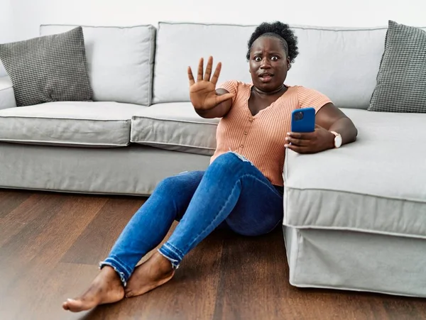 年轻的非洲女人用智能手机坐在家里的地板上 手牵着手 用愤怒和沮丧的表情做着停下来的手势 — 图库照片