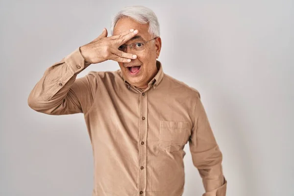 Hispanic senior man wearing glasses peeking in shock covering face and eyes with hand, looking through fingers with embarrassed expression.