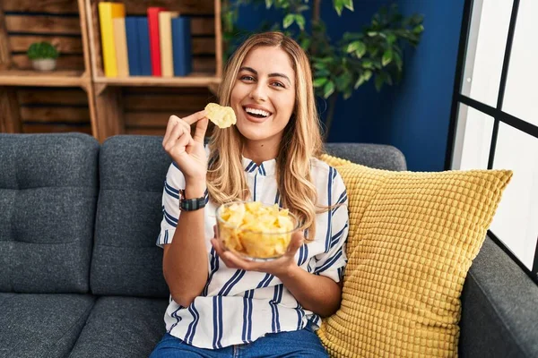 Young Blonde Woman Eating Chips Potatoes Sitting Sofa Home — Stok fotoğraf