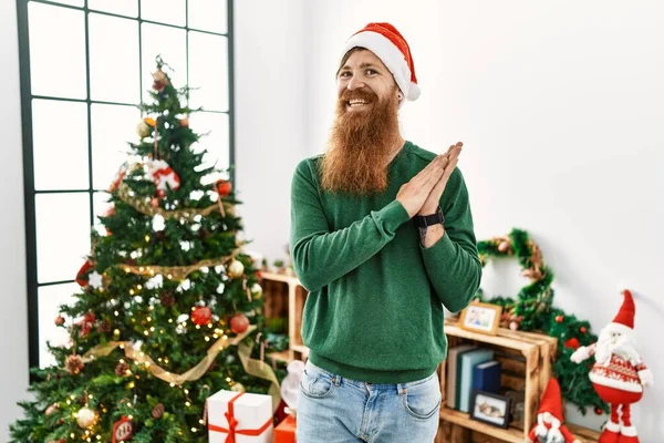 Redhead man with long beard wearing christmas hat by christmas tree clapping and applauding happy and joyful, smiling proud hands together