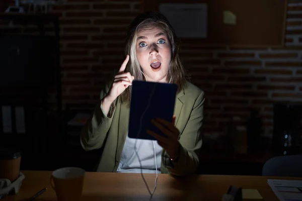 Blonde caucasian woman working at the office at night smiling amazed and surprised and pointing up with fingers and raised arms.