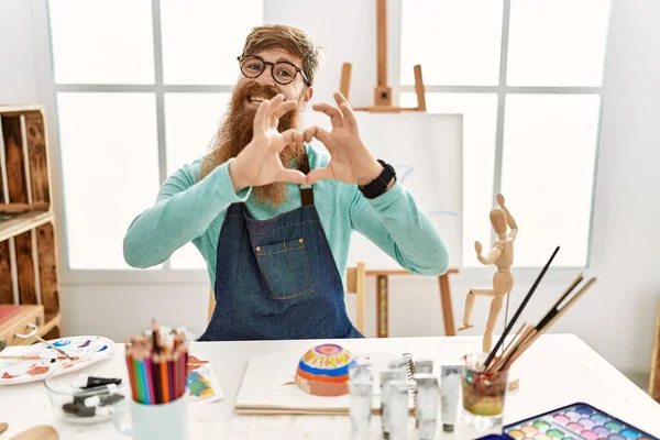 Redhead man with long beard painting clay bowl at art studio smiling in love doing heart symbol shape with hands. romantic concept.