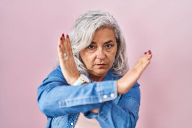 Middle age woman with grey hair standing over pink background rejection expression crossing arms doing negative sign, angry face 