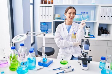 Young brunette woman working at scientist laboratory smiling happy pointing with hand and finger to the side 