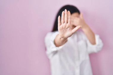 Young brunette woman standing over pink background covering eyes with hands and doing stop gesture with sad and fear expression. embarrassed and negative concept. 