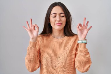 Genç esmer kadın beyaz arka planda duruyor rahatlamış ve gözleri kapalı gülümsüyor parmaklarıyla meditasyon hareketi yapıyor. yoga konsepti. 
