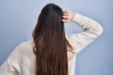 Genç esmer kadın mavi arka planda geriye doğru duruyor. Elleri başının üstünde şüpheyi düşünüyor. 