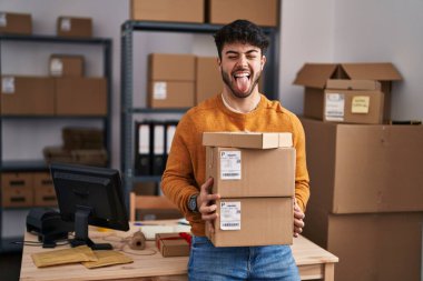 Hispanic man with beard working at small business ecommerce holding packages sticking tongue out happy with funny expression. 