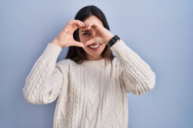 Genç esmer kadın mavi arka planda duruyor. El ve parmaklarıyla kalp şeklinde, tabelanın içinden bakıp gülümsüyor. 