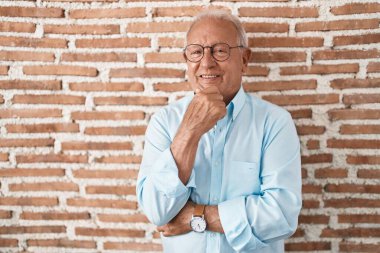 Senior man with grey hair standing over bricks wall looking confident at the camera smiling with crossed arms and hand raised on chin. thinking positive. 