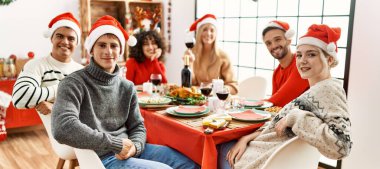 Bir grup genç arkadaş Noel yemeği yiyor. Masada oturan kameraya bakıyorlar..