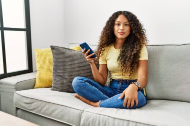 Evdeki kanepede oturan güzel İspanyol kadın akıllı telefon kullanıyor. Yüzünde ciddi bir ifade var. Basit ve doğal bir şekilde kameraya bakıyorsun.. 