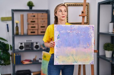 Genç sarışın kadın tuvali tutuyor gülümsüyor yan tarafa bakıyor ve düşünüyor.. 