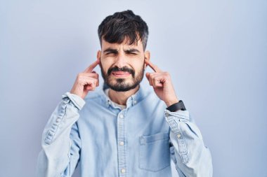 Young hispanic man with beard standing over blue background covering ears with fingers with annoyed expression for the noise of loud music. deaf concept. 