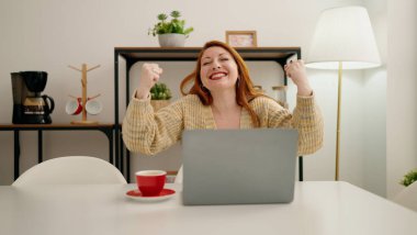 Young redhead woman using laptop with celebrate expression at home