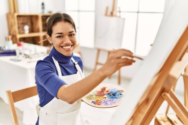 Sanat stüdyosunda kendine güvenen genç Latin kadın gülümsüyor.