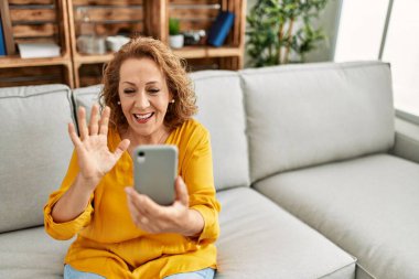 Orta yaşlı beyaz bir kadın evdeki kanepede otururken akıllı telefon kullanıyor..