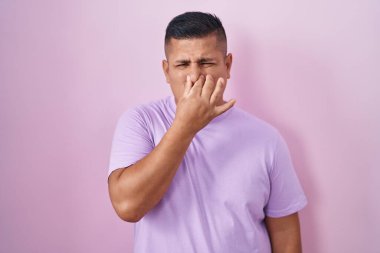 Young hispanic man standing over pink background smelling something stinky and disgusting, intolerable smell, holding breath with fingers on nose. bad smell 