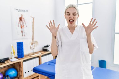 Young caucasian woman working at pain recovery clinic celebrating mad and crazy for success with arms raised and closed eyes screaming excited. winner concept 