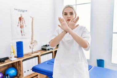 Young caucasian woman working at pain recovery clinic rejection expression crossing arms and palms doing negative sign, angry face 