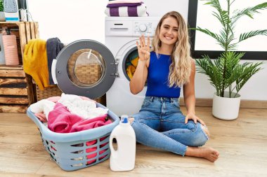 Young beautiful woman doing laundry sitting by wicker basket showing and pointing up with fingers number four while smiling confident and happy. 