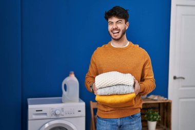 Hispanic man with beard holding clean folded laundry angry and mad screaming frustrated and furious, shouting with anger. rage and aggressive concept. 