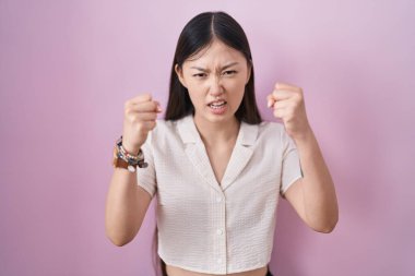 Pembe arka planda duran Çinli genç bir kadın öfkeyle bağırırken kızgın ve kızgın yumruklarını kaldırıyor. öfke ve agresif konsept. 