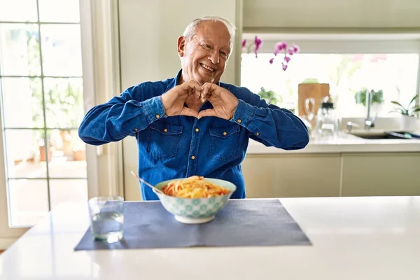 Senior man with grey hair eating pasta spaghetti at home smiling in love doing heart symbol shape with hands. romantic concept.