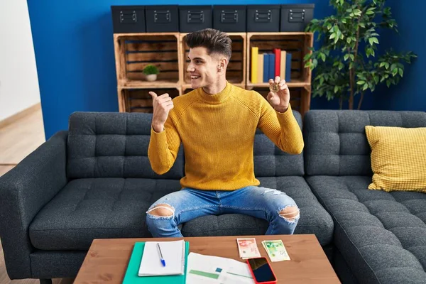 Young hispanic man holding bitcoin virtual money pointing thumb up to the side smiling happy with open mouth 