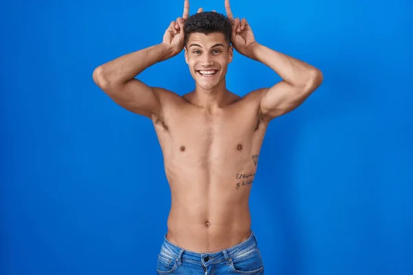 Young hispanic man standing shirtless over blue background posing funny and crazy with fingers on head as bunny ears, smiling cheerful