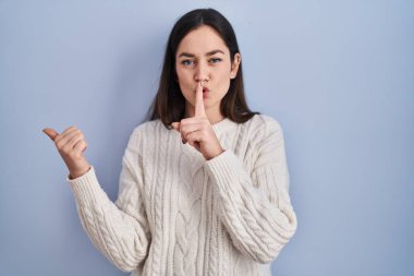 Genç esmer kadın mavi arka planda dikiliyor ve sessiz olmak istiyor. Parmakları dudaklarında, elleri yandan işaret ederek. sessizlik ve gizli konsept. 