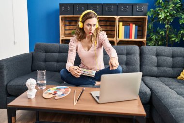 Young beautiful woman practicing painting looking at online tutorial pointing with finger to the camera and to you, confident gesture looking serious 