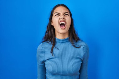 Mavi arka plan üzerinde duran genç Brezilyalı kadın öfkeli ve öfkeli çığlık atıyor öfkeyle bağırıyor. öfke ve agresif konsept. 