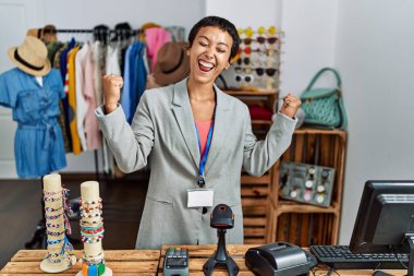 Perakende mağazasında yönetici olarak çalışan kısa saçlı genç İspanyol kadın kolları havada, gülümseyen ve başarı çığlıkları atan, çok mutlu ve heyecanlı bir jest yapıyor. kutlama konsepti. 
