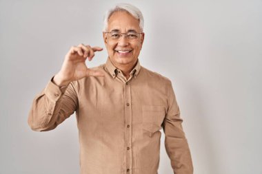 Hispanic senior man wearing glasses smiling and confident gesturing with hand doing small size sign with fingers looking and the camera. measure concept. 