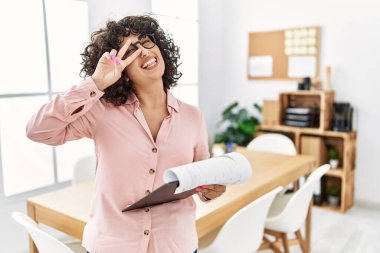 Young middle eastern woman wearing business style at office doing peace symbol with fingers over face, smiling cheerful showing victory 