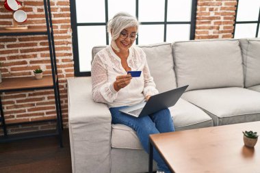 Evdeki koltukta oturan orta yaşlı bir kadın dizüstü bilgisayar ve kredi kartı kullanıyor.