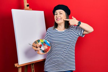 Middle age hispanic woman standing drawing with palette by painter easel stand smiling cheerful showing and pointing with fingers teeth and mouth. dental health concept. 