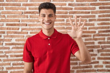 Young hispanic man standing over bricks wall showing and pointing up with fingers number five while smiling confident and happy. 