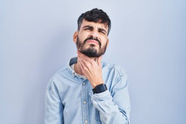 Young hispanic man with beard standing over blue background touching painful neck, sore throat for flu, clod and infection 