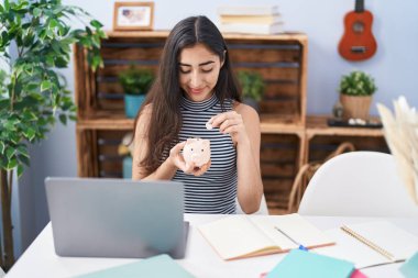 Young hispanic girl smiling confident inserting coin on piggy bank at home