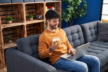 Young hispanic man using laptop and drinking coffee sitting on sofa at home