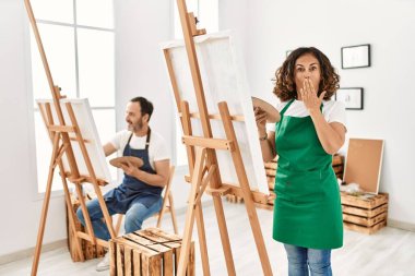 Hispanic middle age woman and mature man at art studio covering mouth with hand, shocked and afraid for mistake. surprised expression 