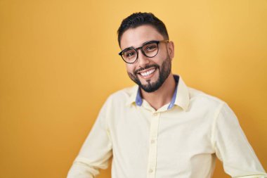 İş kıyafetleri ve gözlük takan İspanyol genç adam, güler yüzlü ve kollarını açıp gülümsüyor. Hoş geldin, olumlu ve kendinden emin selamlar. 