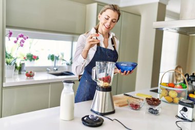 Genç sarışın kadın mutfaktaki blendere buz dökerek gülümsüyor.