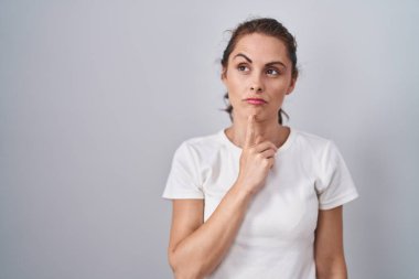 Beautiful brunette woman standing over isolated background thinking concentrated about doubt with finger on chin and looking up wondering 