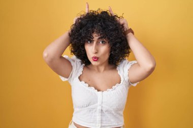 Young brunette woman with curly hair standing over yellow background doing funny gesture with finger over head as bull horns 