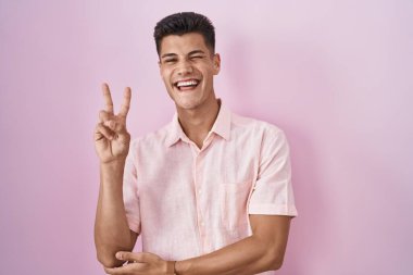 Young hispanic man standing over pink background smiling with happy face winking at the camera doing victory sign with fingers. number two. 